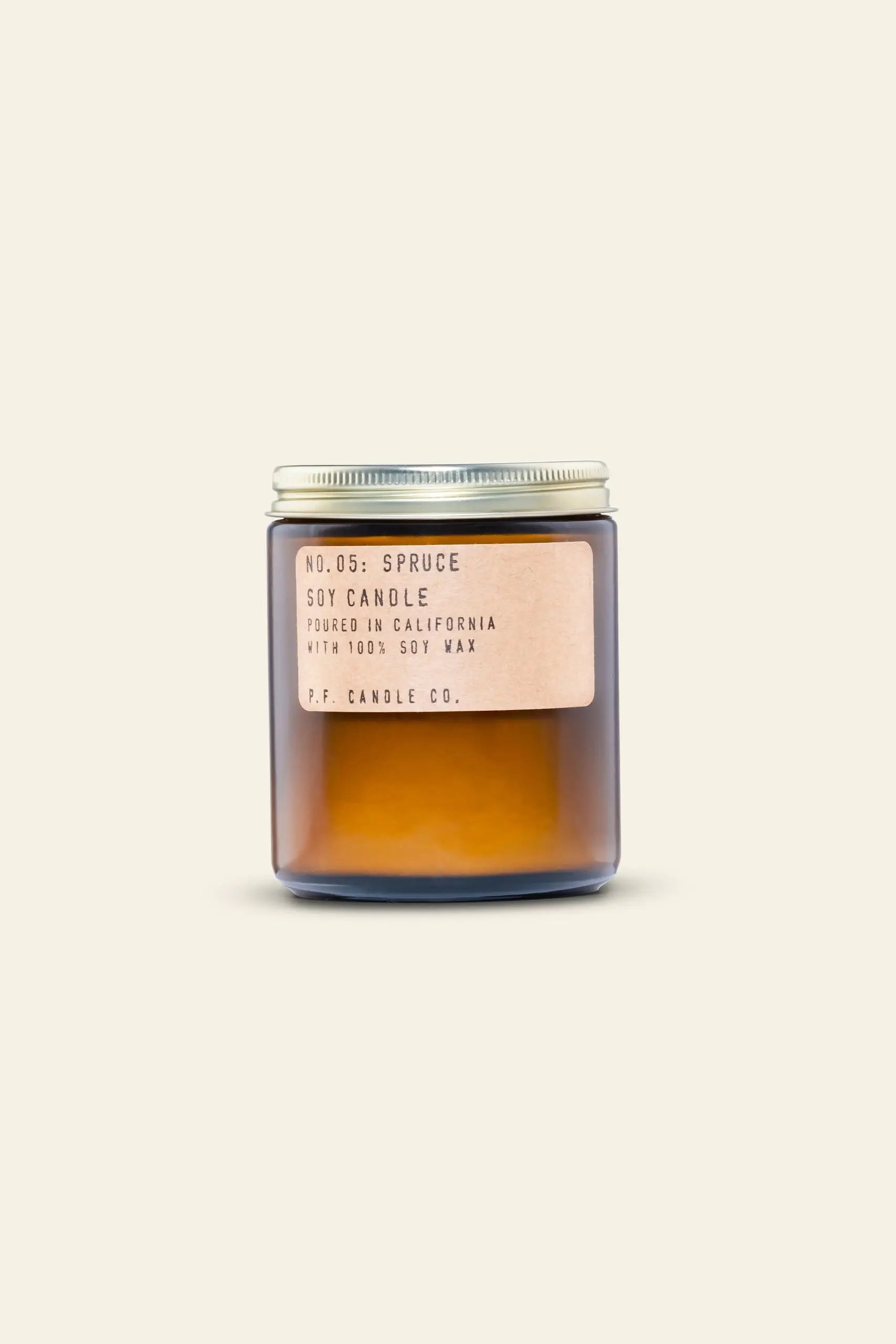 A brown glass jar with a metal lid holds a P.F. Candle Co. Spruce 7.2 oz Soy Candle made from 100% soy wax, poured in California. The label features the brand and scent details. The background is off-white.