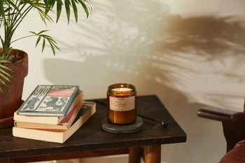 The P.F. Candle Co. Spruce 7.2 oz Soy Candle glows atop a round coaster beside books on a wooden table, its Blue Spruce scent filling the air as soft shadows from a potted plant and nearby chair dance on the cream-colored wall.