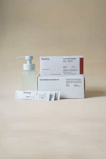 A Pass It On 0.02 Refillable Hand Wash Set (Pack of 3-Experiential Bundle) by Pass It On, featuring a clear Perpetual Bottle, two minimalist white boxes with black text and red accents, and two sachets, is arranged on a light beige surface.
