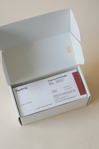 A white cardboard box lies open on a light surface, revealing a Pass It On 0.02 Refillable Hand Wash Set, Pack of 3-Experiential Bundle by Pass It On, with an informational card and sustainable botanically-derived powder inside.
