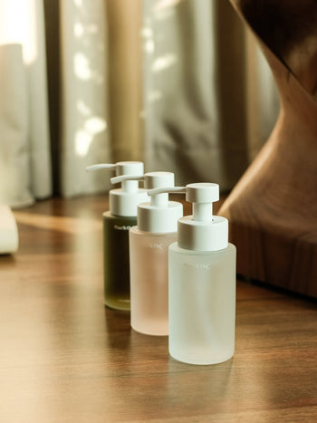 Three Pass It On 0.02 Refillable Hand Wash bottles from the Pack of 3-Experiential Bundle, each with a white lid and botanically-derived powder in various hues, sit on wood as sunlight streams through curtains behind.
