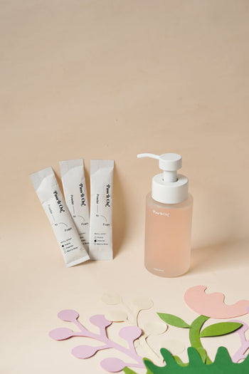 The Pass It On 0.02 Refillable Hand Wash Set, Pack of 3-Oleander features a peach liquid pump bottle beside three white sachets, all by Pass It On, artfully arranged on a neutral backdrop with pastel paper cutouts for a sustainable vibe.