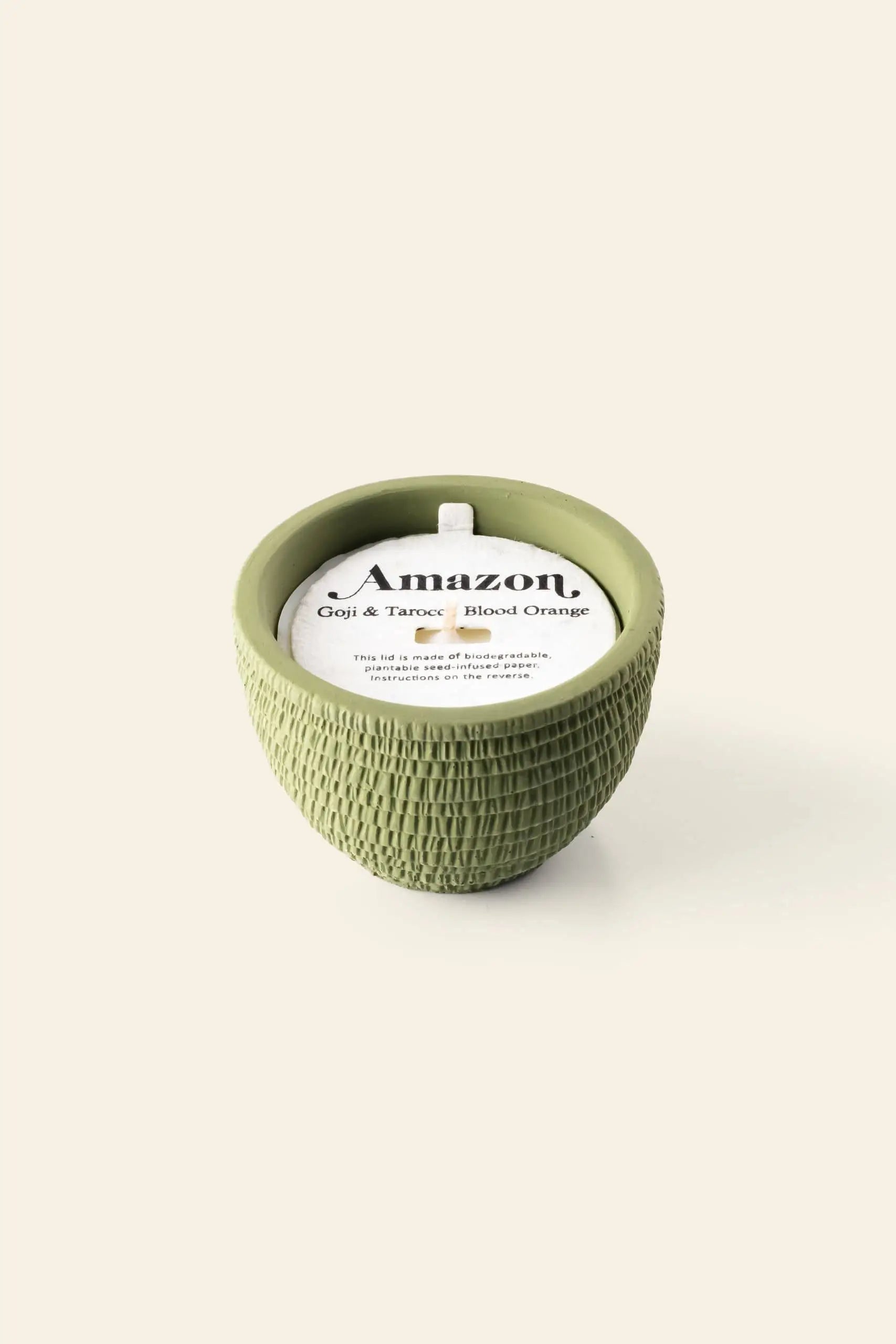 A green textured Pass It On Amazon Candle by Pass It On, labeled Goji & Tarocco Blood Orange, rests on a light beige background, radiating exotic fruity fragrances.