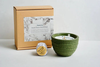 The Pass It On Amazon Candle, a green textured bowl, sits beside a small round wax melt and a tan Goji & Tobacco Blood Orange box with botanical label—all by Pass It On—displaying exotic fragrances on a white surface.