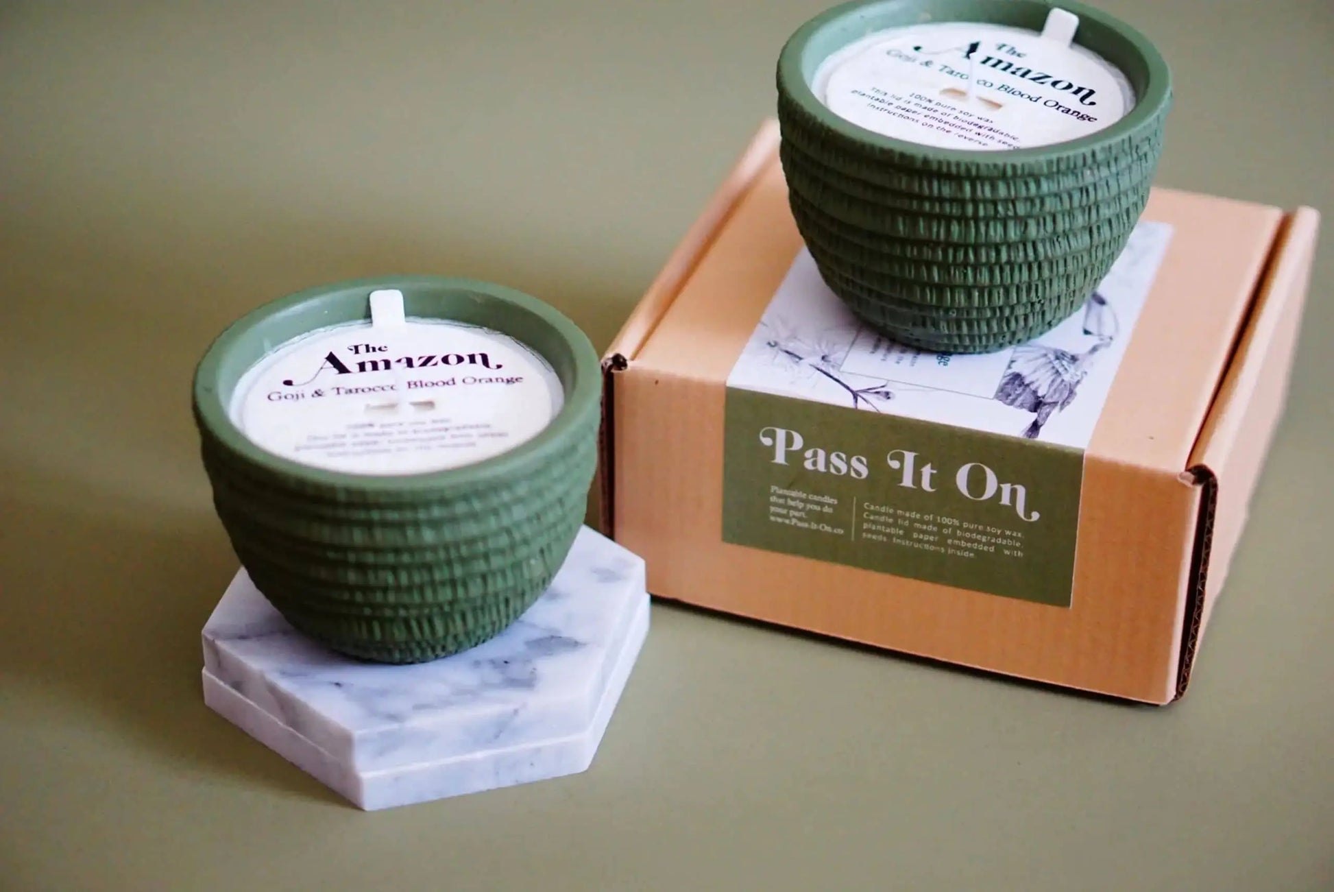 Two Pass It On Amazon Candles with green textured glass and white labels, featuring exotic scents, rest on a hexagonal marble coaster and a Pass It On box against a muted green background.