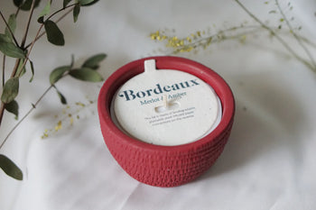 A round red Pass It On Bordeaux Candle with a textured exterior sits on a white surface amid greenery. The candle, labeled with white, blue, and gold text, evokes rich red wine and black cherry scents. Brand: Pass It On.