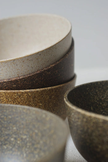 A close-up of stacked Pass It On Capsule Bowls, crafted from upcycled rice husks in earthy beige, brown, and speckled dark gray tones, showcases the brands sustainable design against a soft, neutral background.