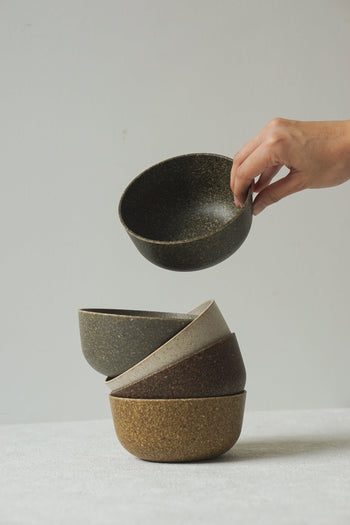 A hand lifts the Pass It On Capsule Bowl, Rice Husk—made from upcycled rice husks—from a stack of four earthy bowls on a light textured surface, set against a plain white background.