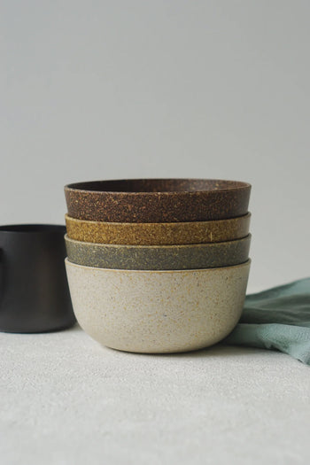 A stack of four Pass It On Capsule Bowls, Rice Husk, in varied earth tones from light to dark, crafted sustainably by Pass It On, sits beside a black cup and light blue cloth on a textured white surface.