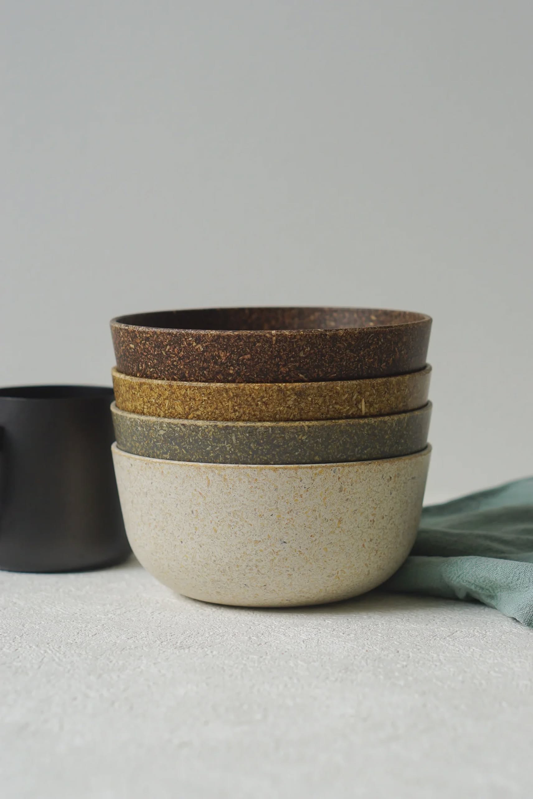 A stack of four Pass It On Capsule Bowls, made from rice husk in natural earth tones, rests on a textured surface beside a black cup and green cloth—ideal for those who value sustainability and mindful design.