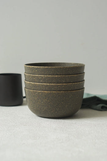 Four stacked, speckled brown Pass It On Capsule Bowls made from upcycled rice husks rest on a light surface, with a dark cup and green cloth in the background—showcasing sustainable style by Pass It On.