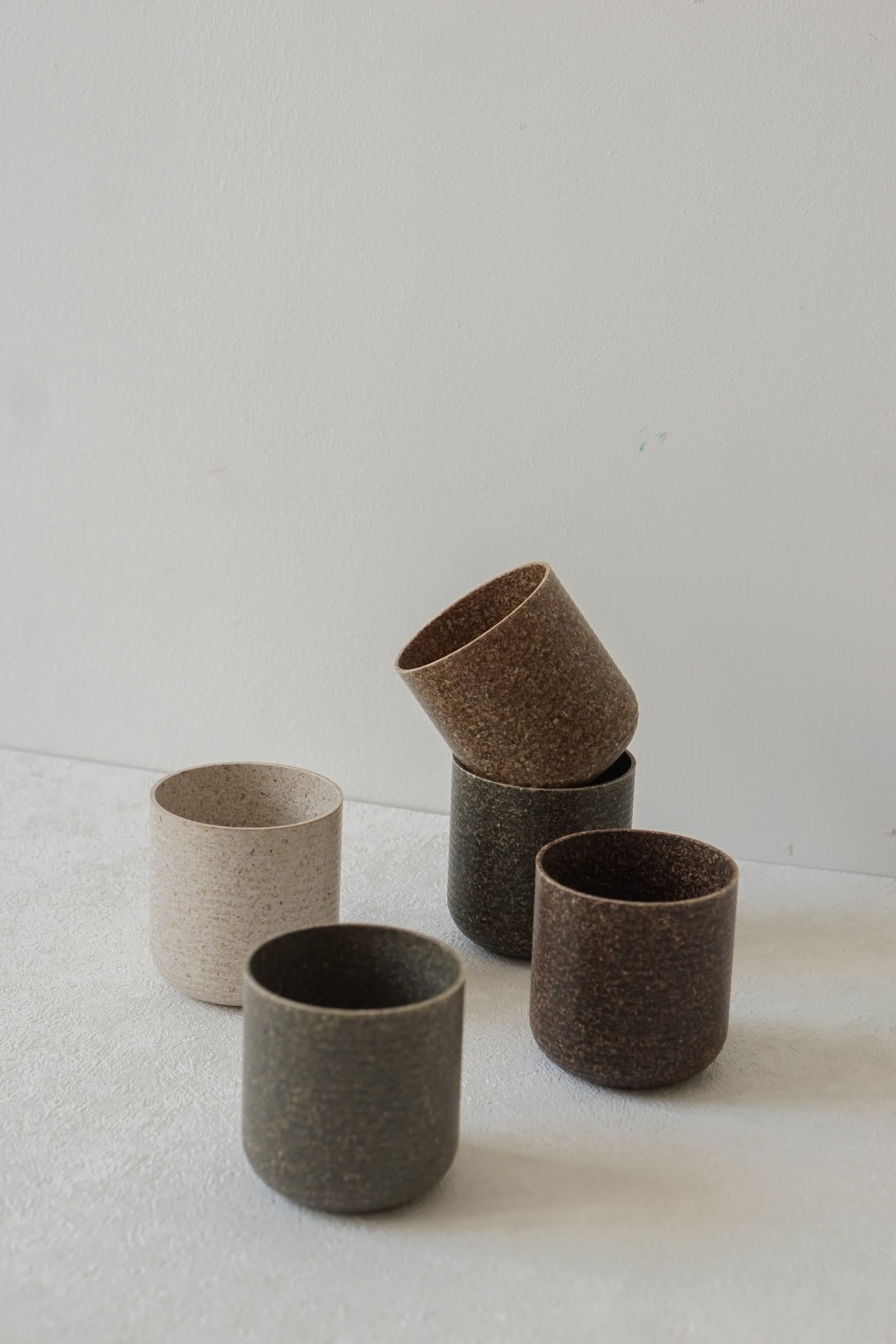 Five minimalist Pass It On Capsule Cups, made from upcycled rice husk by Pass It On, are displayed in earthy tones—two stacks of two on a light surface and one cup in the foreground—for an eco-conscious alternative.