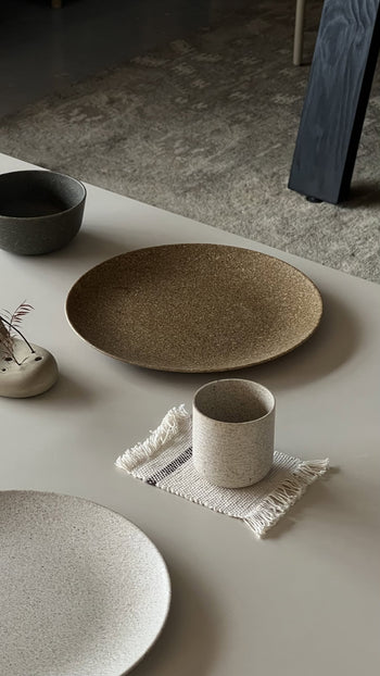 A minimalist table setting featuring Pass It On’s Dining Plate L in Rice Husk, a beige cup on a fringed cloth, a small gray bowl, and a ceramic vase with dried flowers—all arranged on a light surface for an eco-conscious look.