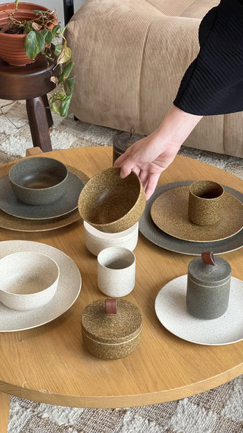 A person’s hand picks up a Pass It On Dining Plate L, Rice Husk from a round wooden table set with assorted eco-conscious tableware in earthy tones. A potted plant and beige furniture can be seen in the background.
