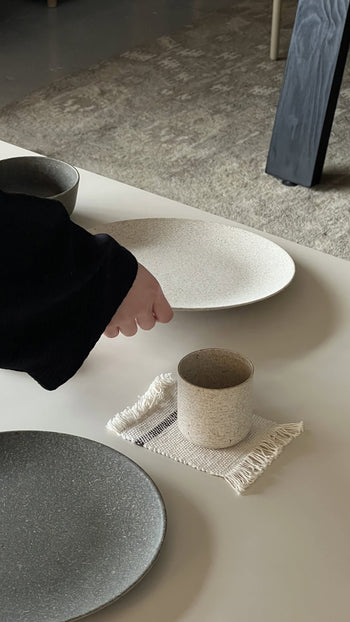 A hand reaches for the Pass It On Dining Plate L, Rice Husk by Pass It On—a speckled white eco-friendly ceramic plate—on a table set with sustainable tableware atop a fringed beige mat, with carpet and table leg in view.