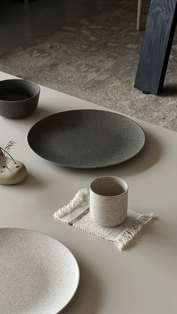 A minimalist tabletop features the Pass It On Dining Plate L made from rice husk, neutral-toned, paired with a cup on a fringed cloth, eco-conscious tableware, and a small vase with dried foliage on a light-colored surface.