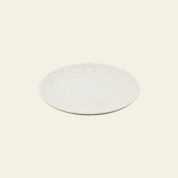 The Pass It On Dining Plate M, Rice Husk by Pass It On is a round, off-white ceramic plate with a subtly textured surface and modern design, providing an eco-friendly choice for sustainable dining. Shown on a plain light beige background.