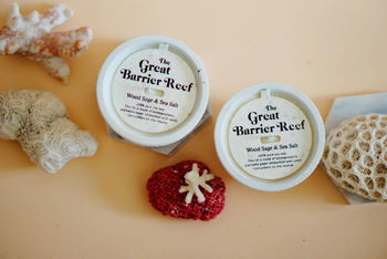 Two round Pass It On Great Barrier Reef Candles sit on a peach surface amid coral and a red sea sponge, capturing the fresh, mineral salt scent of the ocean—ideal for aromatherapy.