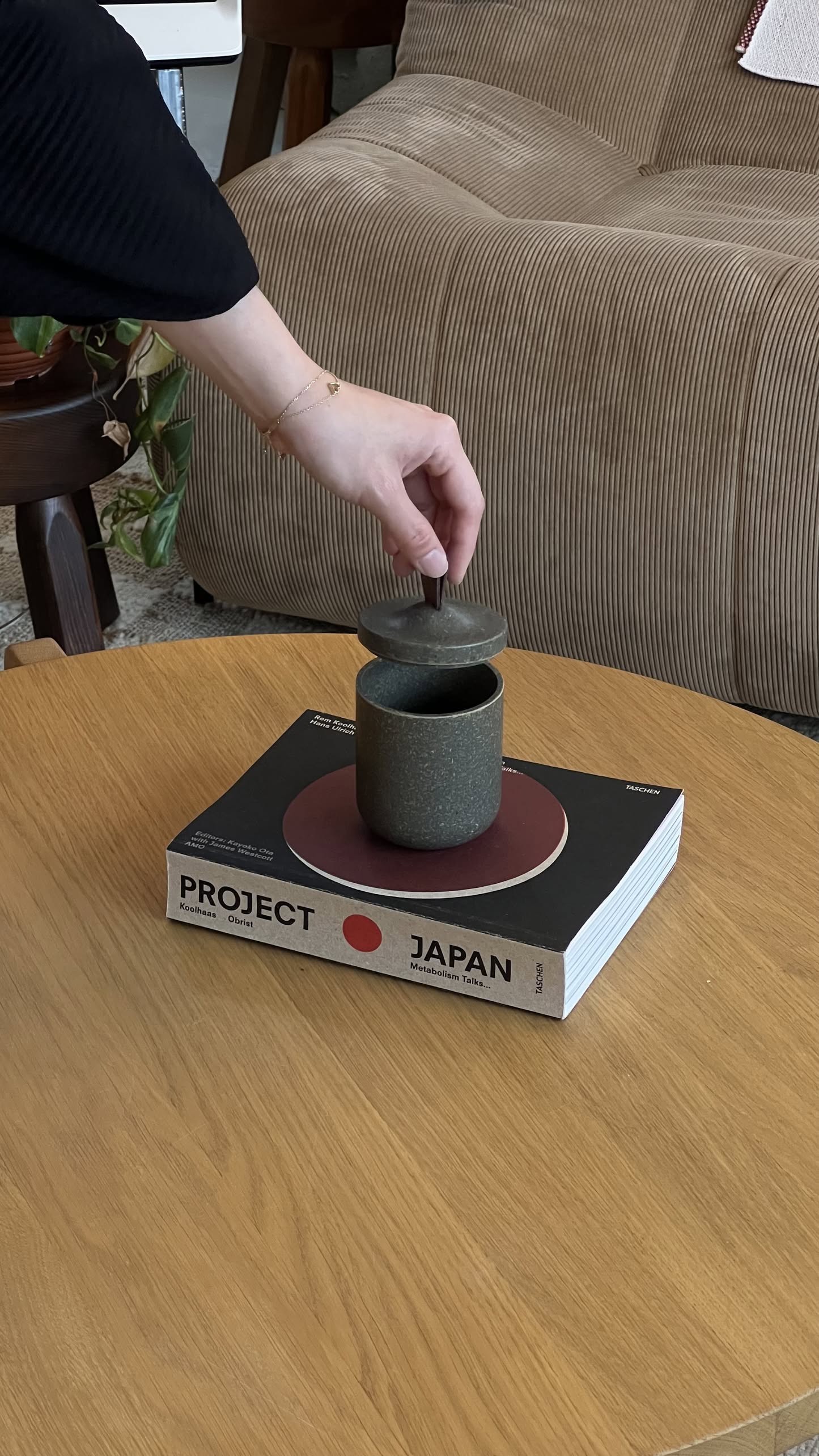 A hand lifts the lid of a Pass It On Tagine Tall Box, Rice Husk by Pass It On, set on a book titled Project Japan atop a wooden table next to a beige sofa.