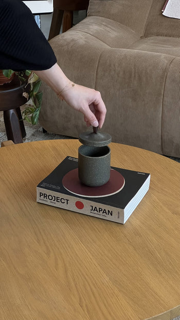 A hand lifts the lid of a Pass It On Tagine Tall Box, Rice Husk by Pass It On, set on a book titled Project Japan atop a wooden table next to a beige sofa.