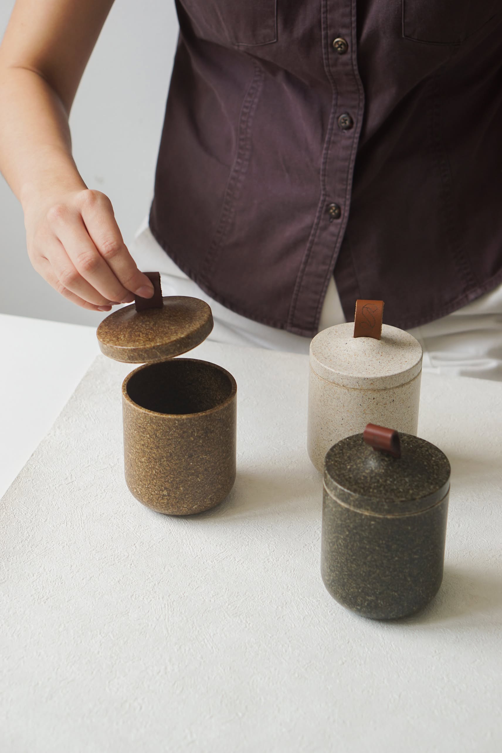 A person in a purple shirt opens the lid of a brown Pass It On Tagine Tall Box, Rice Husk, with two other lidded containers—one white and one dark green—on an eco-friendly white surface.
