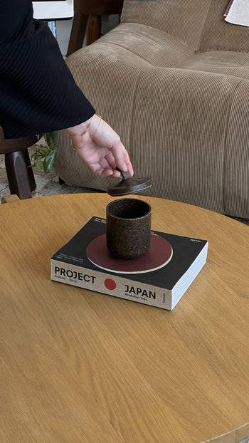A hand holds a spoon above a dark cup next to the Pass It On Tagine Tall Box, Rice Husk—a chic, upcycled piece from Pass It On—on a wooden coffee table in front of a beige sofa.