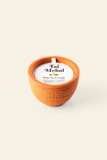 The Pass It On Taj Mahal Candle features a round, textured terracotta holder with a white candle inside. The label highlights White Tea & Ginger, offering an exotic aroma inspired by Indian culture. The background is plain and light-colored.