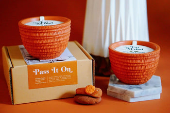 The Pass It On Taj Mahal Candle, known for its exotic aroma, rests on a Pass It On box and marble coaster. A stack of cookies and a white vase add charm, all set on an orange surface inspired by Indian culture.