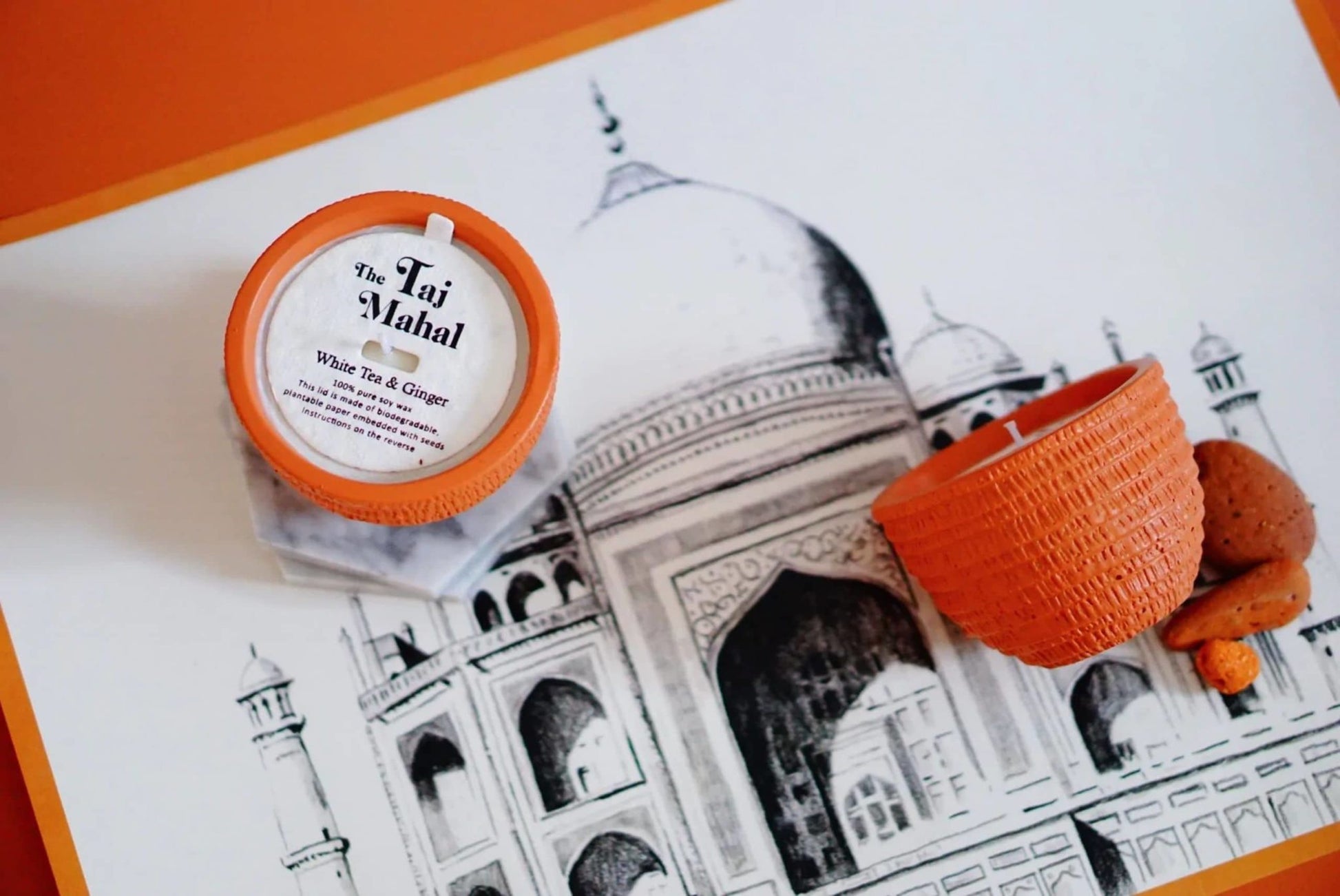 A Pass It On Taj Mahal Candle and an orange textured holder sit on a Taj Mahal drawing, evoking Indian culture. Two brown cookies complete the scene, filled with warm orange accents and an exotic aroma.
