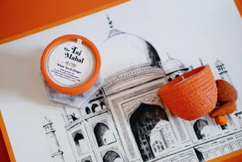 A Pass It On Taj Mahal Candle and an orange textured holder sit on a Taj Mahal drawing, evoking Indian culture. Two brown cookies complete the scene, filled with warm orange accents and an exotic aroma.