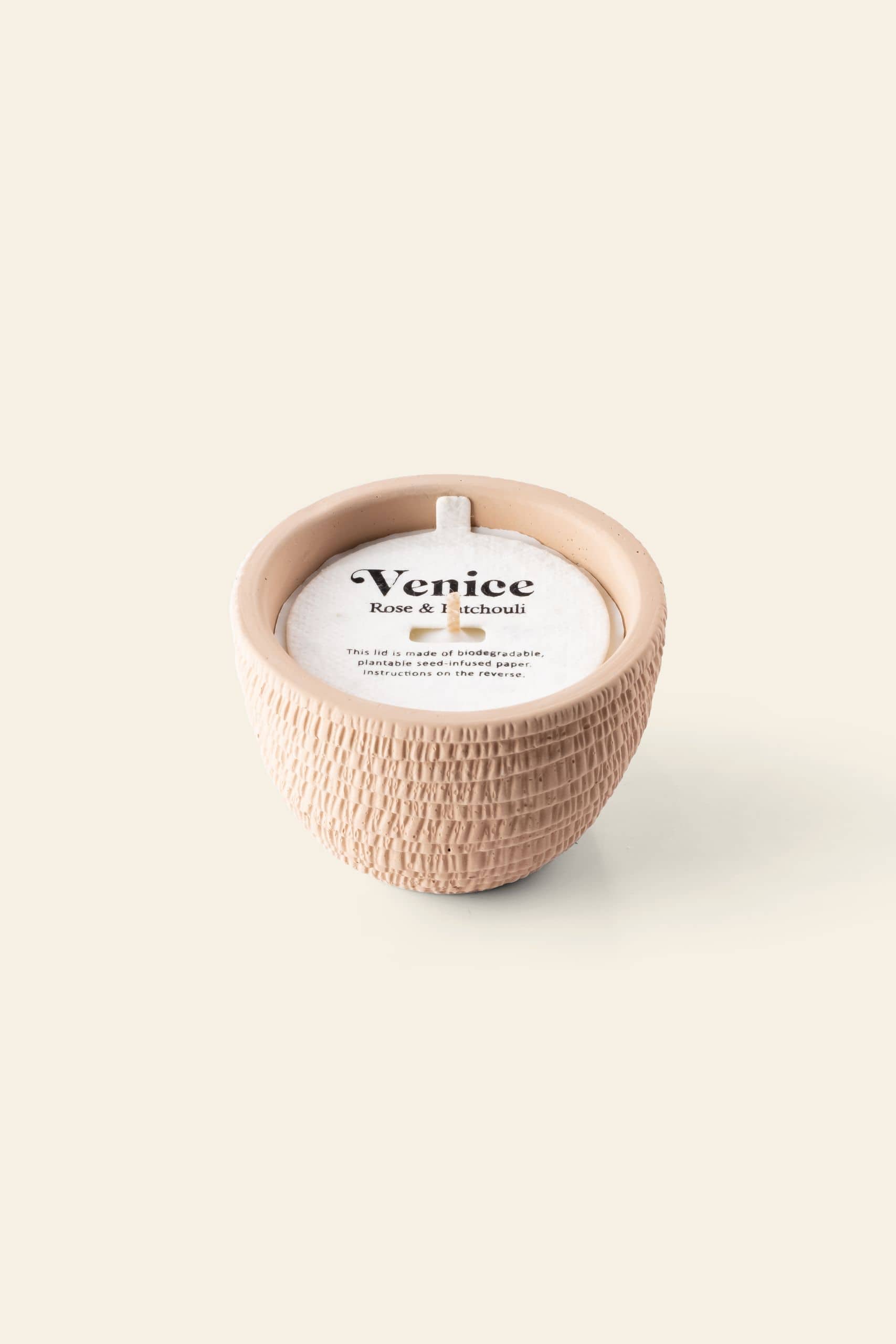 The Pass It On Venice Candle by Pass It On features a textured, round beige ceramic pot, white wax with a single wick, and a label reading “Venice Rose & Patchouli,” evoking romantic rose and patchouli scents. Set against a light beige background.