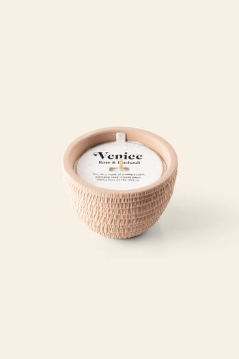 The Pass It On Venice Candle by Pass It On features a textured, round beige ceramic pot, white wax with a single wick, and a label reading “Venice Rose & Patchouli,” evoking romantic rose and patchouli scents. Set against a light beige background.
