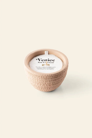 The Pass It On Venice Candle features a textured beige ceramic holder with a white candle, filling your space with romantic rose and patchouli scent. The label reads “Venice, Rose & Patchouli”—ideal for home fragrance lovers.