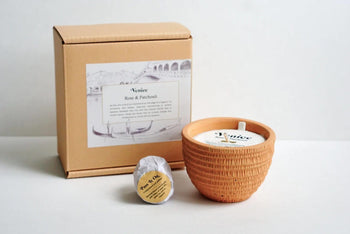 A brown Pass It On box, a small round item with a gold label, and the textured Pass It On Venice Candle are arranged on white. Its illustrated label hints at a patchouli and rose scent for an inviting home fragrance.
