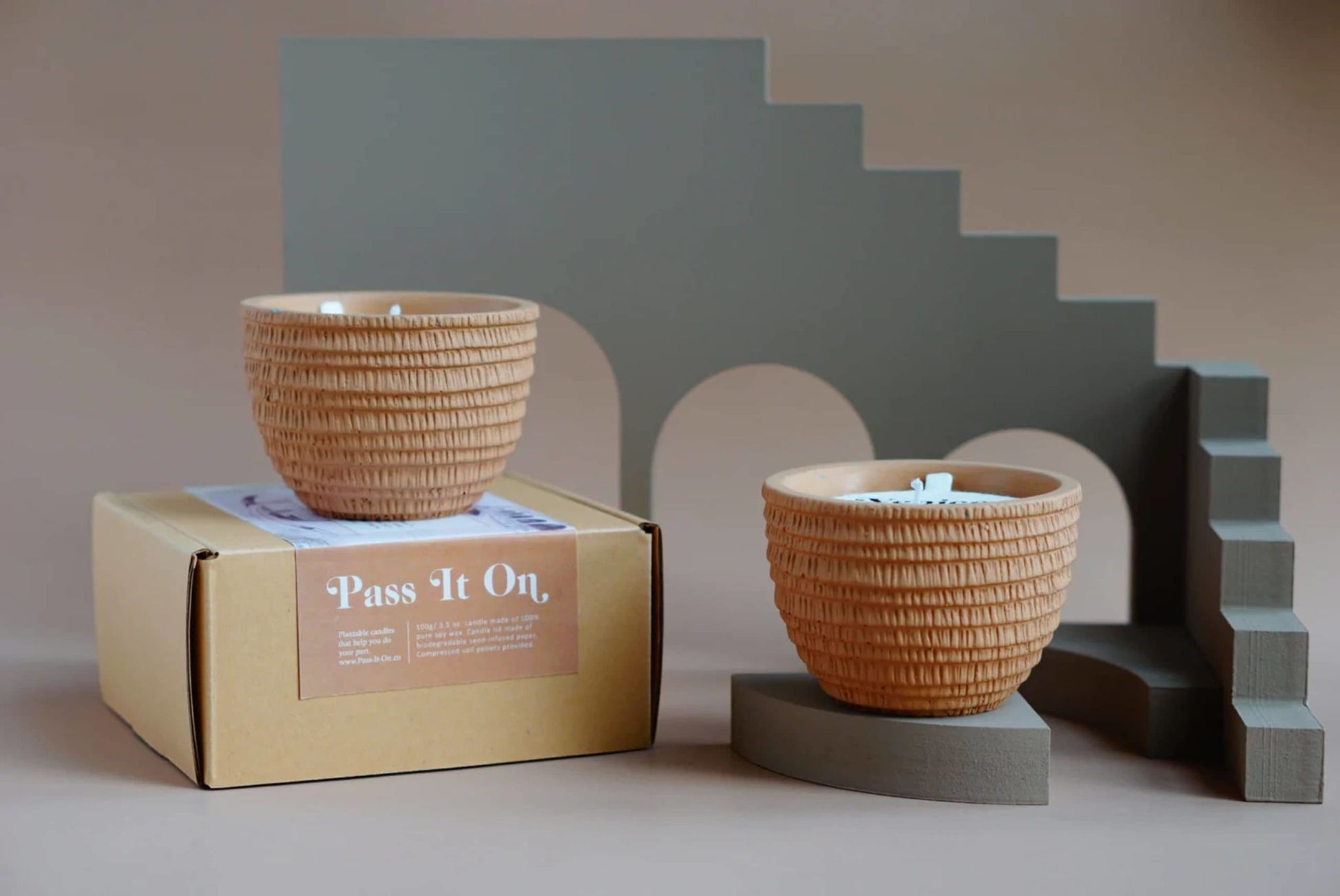 Two textured, light brown Pass It On Venice Candle plant pots sit—one on a box labeled Pass It On, one on a gray stand—with minimalist gray arches and stairs, evoking cozy patchouli and rose notes against a beige background.