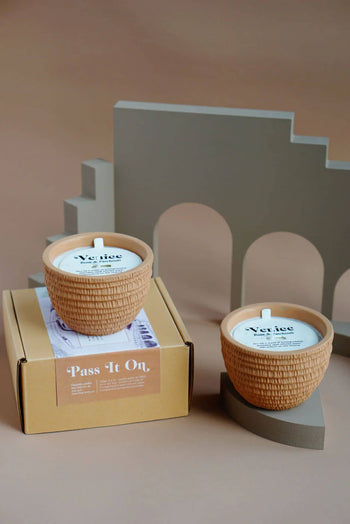 Two textured Pass It On Venice Candles in round tan holders are shown—one on a Pass It On box, the other on a gray surface—filling the room with their patchouli and rose home fragrance.