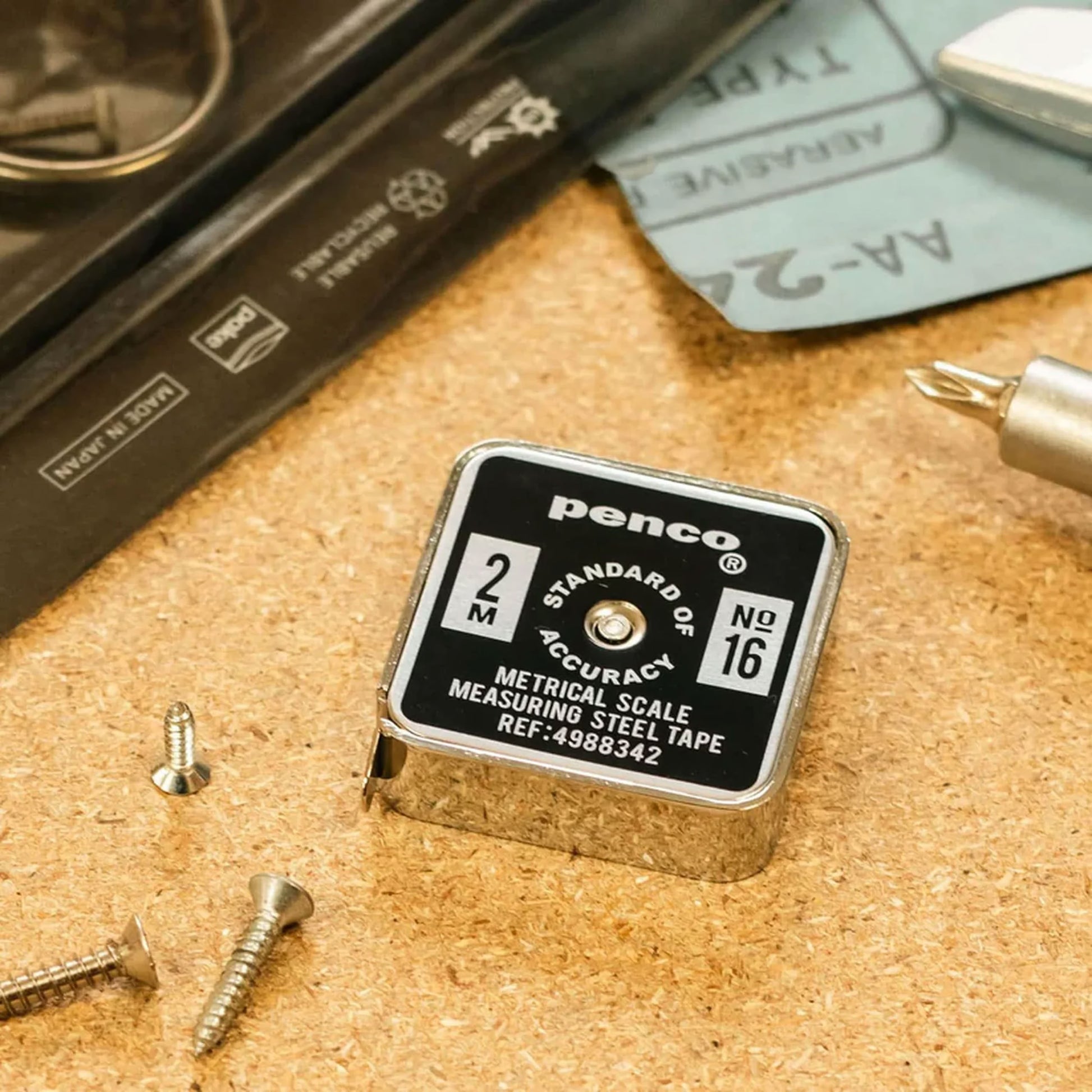 A compact, square Penco Pocket Measure sits on a cork surface beside loose screws, a screwdriver, and a black case. This stainless steel vintage measuring tape by Penco features a retro-inspired label displaying "2M" and "No.16.