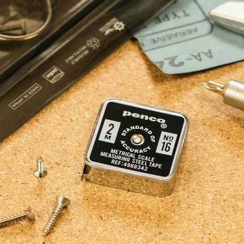 A compact, square Penco Pocket Measure sits on a cork surface beside loose screws, a screwdriver, and a black case. This stainless steel vintage measuring tape by Penco features a retro-inspired label displaying "2M" and "No.16.