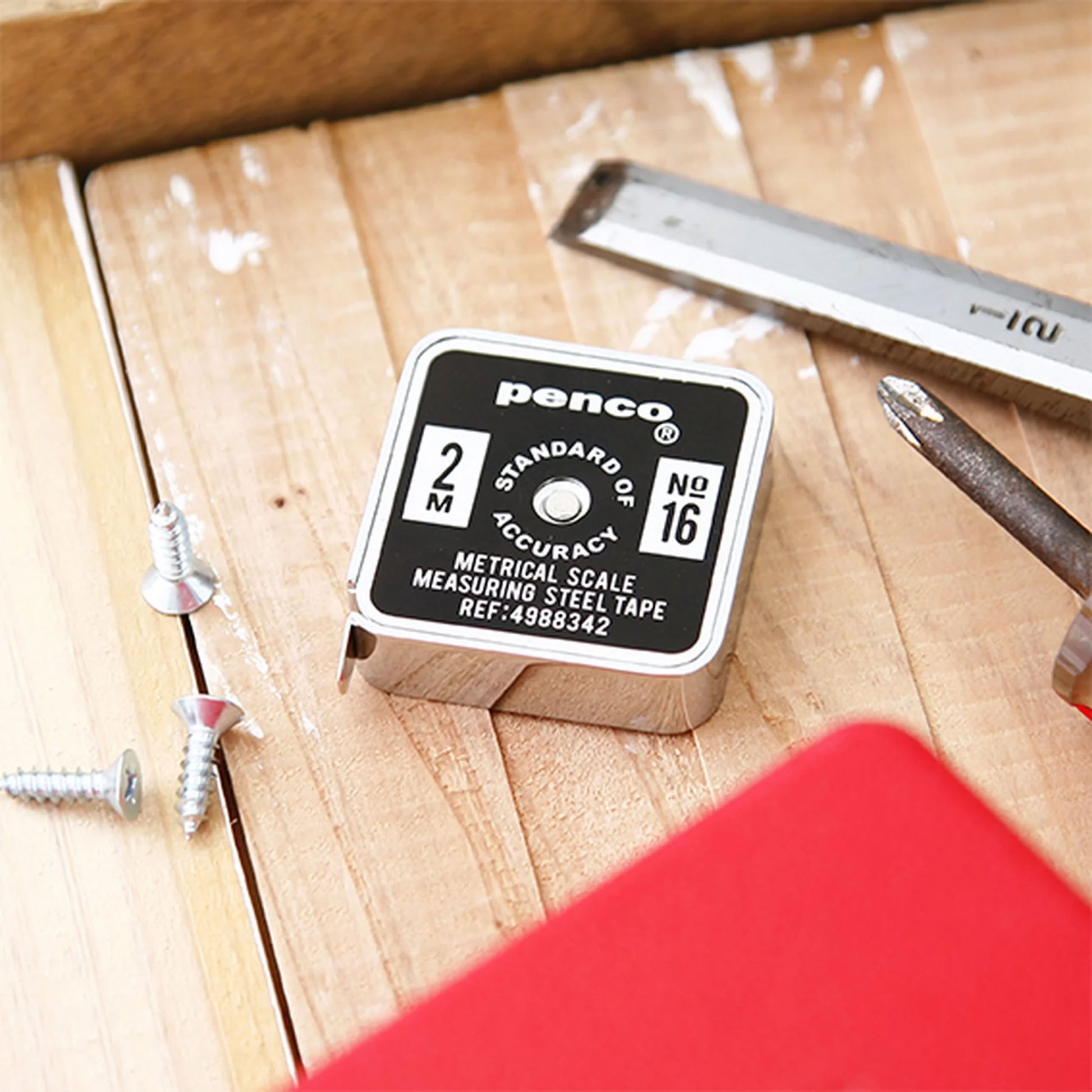 A Penco Pocket Measure, accompanied by screws, a screwdriver, and a square ruler, rests on a wooden surface with part of a red object peeking from the bottom corner, highlighting the retro-inspired aesthetic of the scene.
