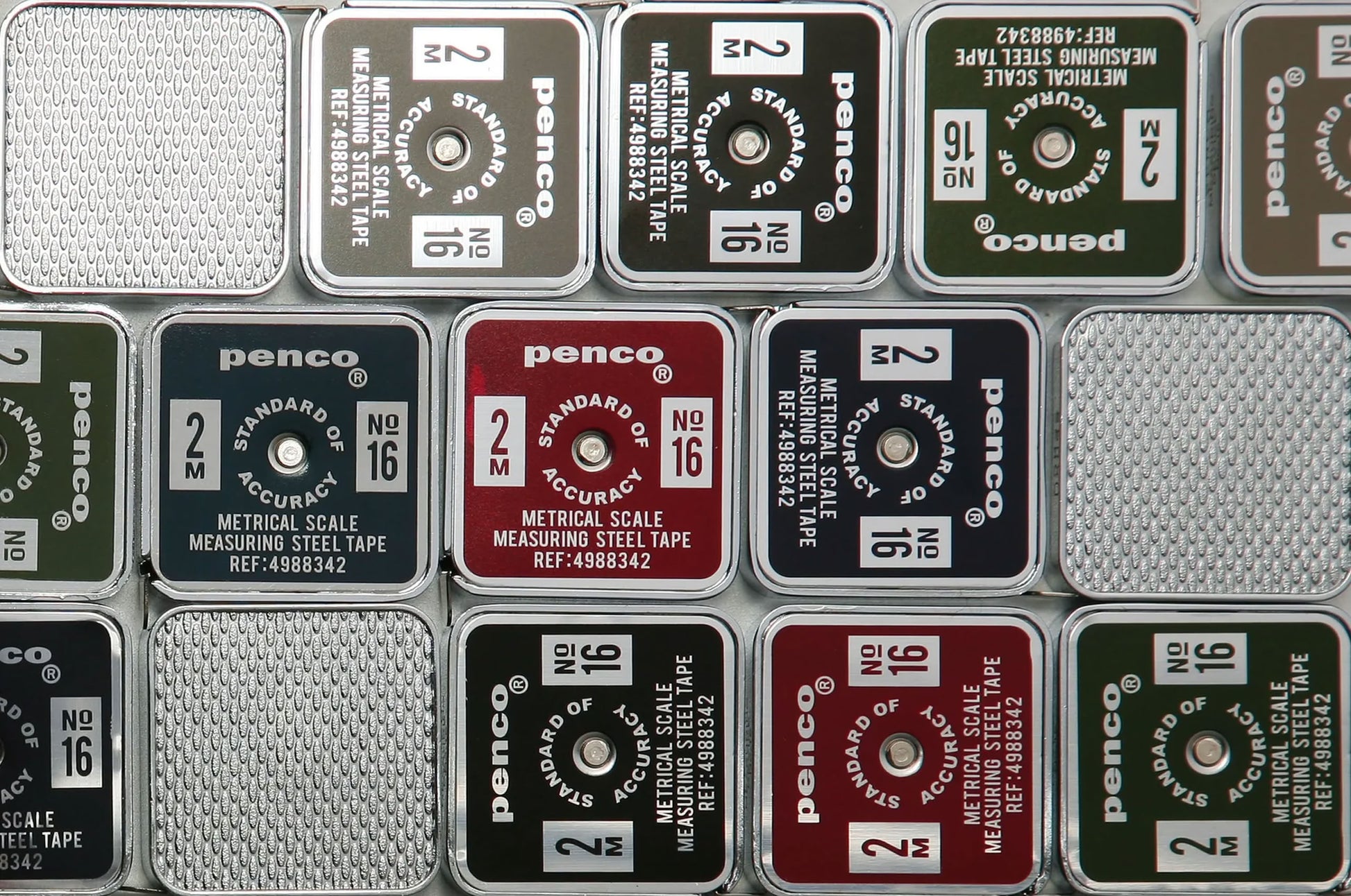Close-up of several stacked, square Penco Pocket Measure cases in red, black, green, and silver, featuring a retro-inspired design with assorted labels and numbers visible on each lid.