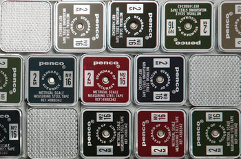 Close-up of several stacked, square Penco Pocket Measure cases in red, black, green, and silver, featuring a retro-inspired design with assorted labels and numbers visible on each lid.