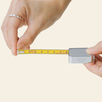 A hand extends the yellow tape of the Penco Pocket Measure—a compact, retro-inspired stainless steel measuring tape by Penco—while another hand holds the end of the tape.