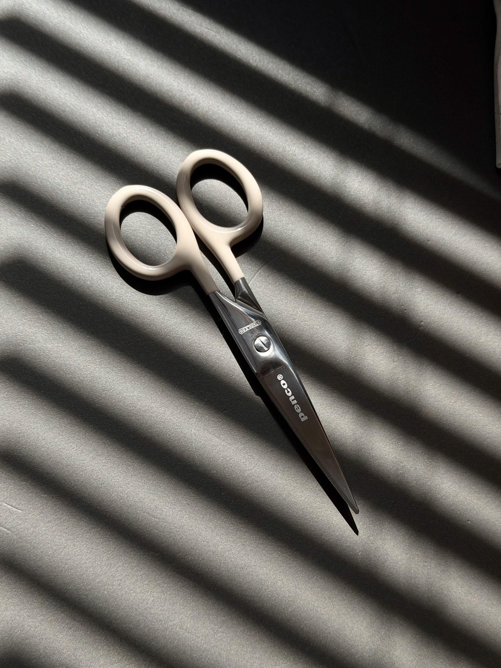 A pair of Penco Stainless Steel Scissors S, featuring beige handles and a subtle retro-industrial design, rests on a dark surface. Sunlight streams through window blinds, casting striped shadows that enhance the classic appeal of this Penco stationery essential.