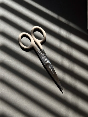 A pair of Penco Stainless Steel Scissors S, featuring beige handles and a subtle retro-industrial design, rests on a dark surface. Sunlight streams through window blinds, casting striped shadows that enhance the classic appeal of this Penco stationery essential.