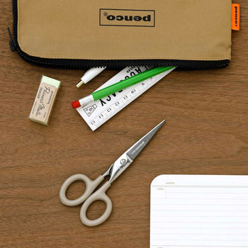A tan pencil case, partially unzipped to reveal a pencil, eraser, white pen, ruler, and tube, sits on a wooden desk alongside the Penco Stainless Steel Scissors S from Penco and a lined notepad—together embodying a timeless stationery classic in retro-industrial style.
