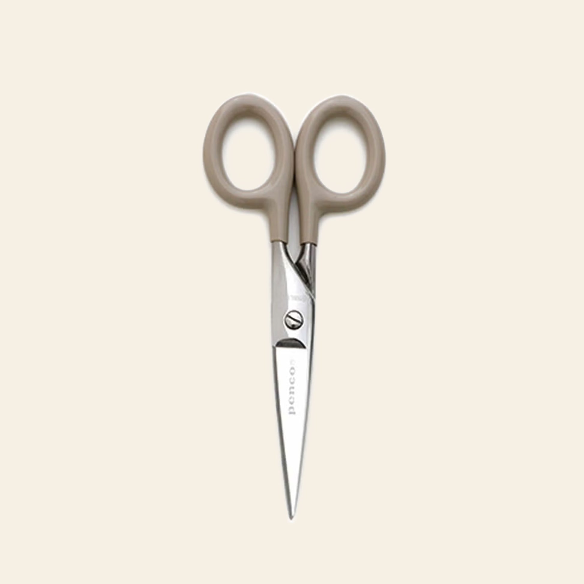 The Penco Stainless Steel Scissors S by Penco, featuring compact stainless steel blades and beige handles, is displayed vertically with the blades pointing downward against a light cream background.