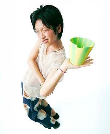 A person with short dark hair winks and smiles, playfully posing with the Plastic Ark Dirty Pot by Plastic Ark—a unique garden pot with green and yellow stripes—while wearing a sheer top, blue tie-dye pants, and beaded accessories against a white background.