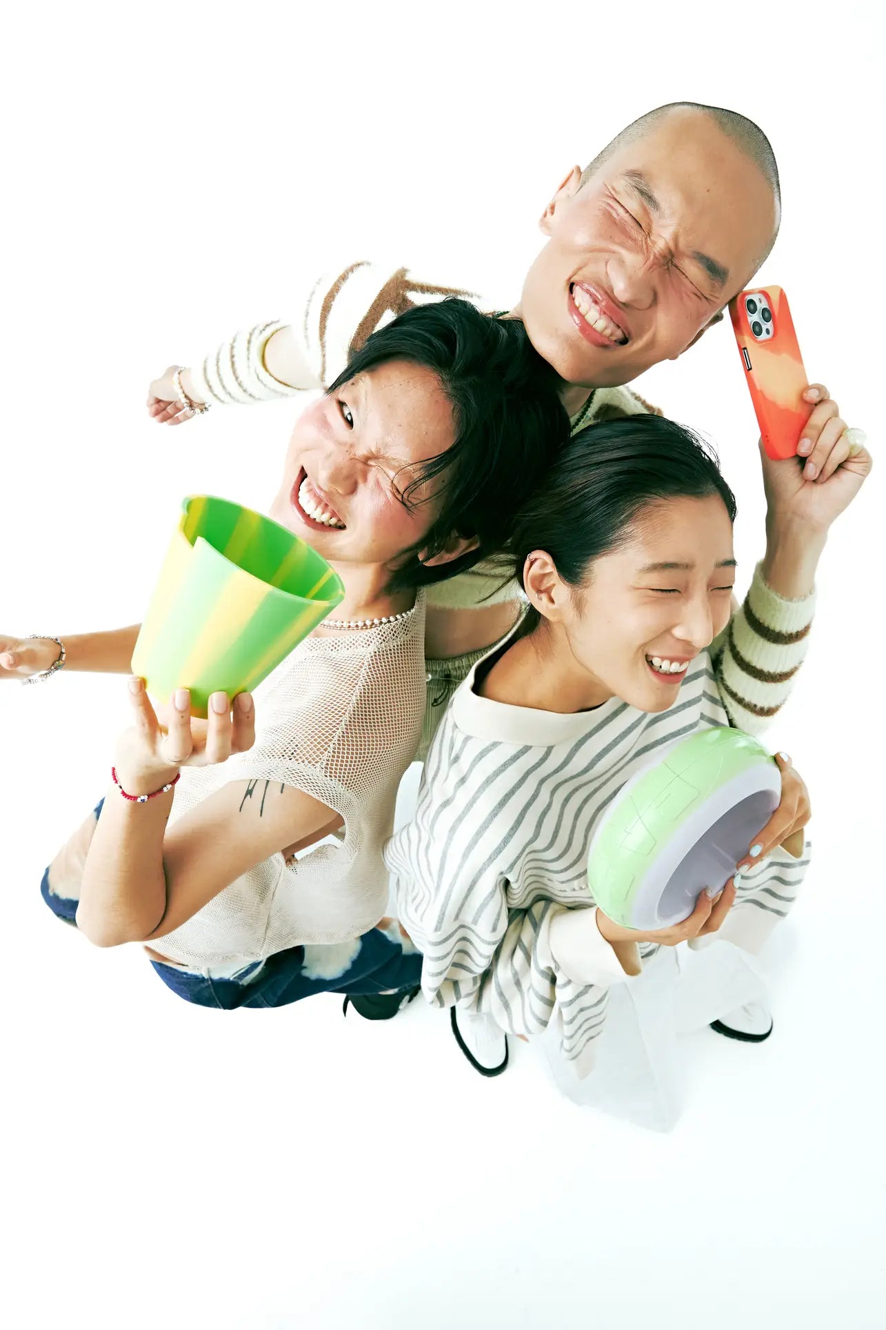 Three people stand close together, smiling and laughing, viewed from above. One holds the Plastic Ark Dirty Pot by Plastic Ark with green-yellow stripes, another a pastel green bowl, and the third an orange smartphone on a white background.