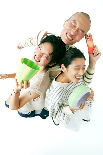 Three people stand close together, smiling and laughing, viewed from above. One holds the Plastic Ark Dirty Pot by Plastic Ark with green-yellow stripes, another a pastel green bowl, and the third an orange smartphone on a white background.
