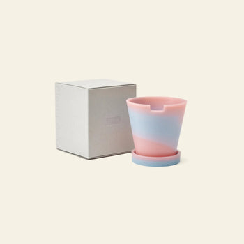 The Plastic Ark Dirty Pot, a pastel pink and light blue marbled plant pot made from recycled plastic with a matching saucer, by Plastic Ark, sits next to a plain light gray box on a beige background.